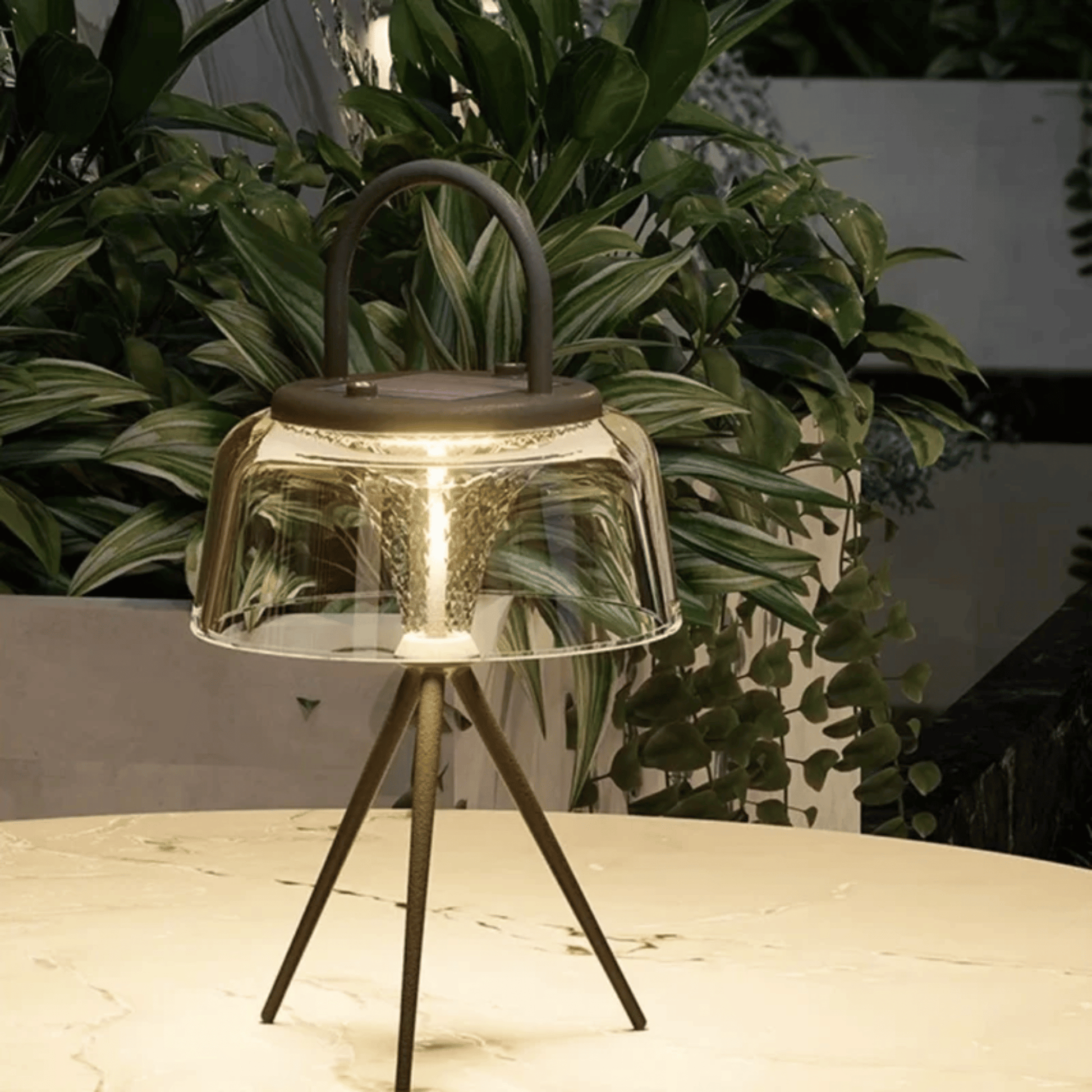 Outdoor lamp with a clear glass shade and metal base on a table with plants in the background