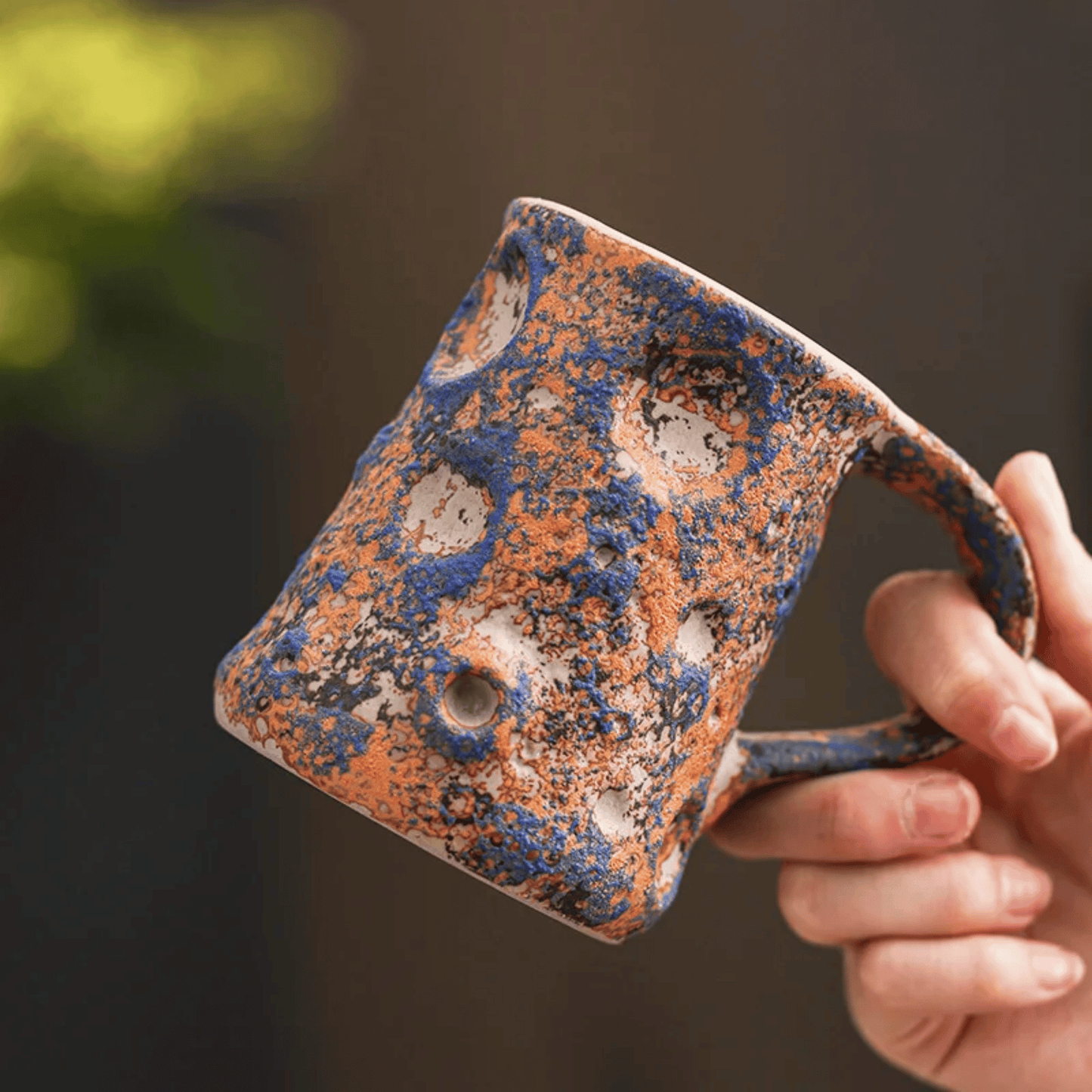 Hand holding a ceramic mug with blue and orange glaze against a blurred background