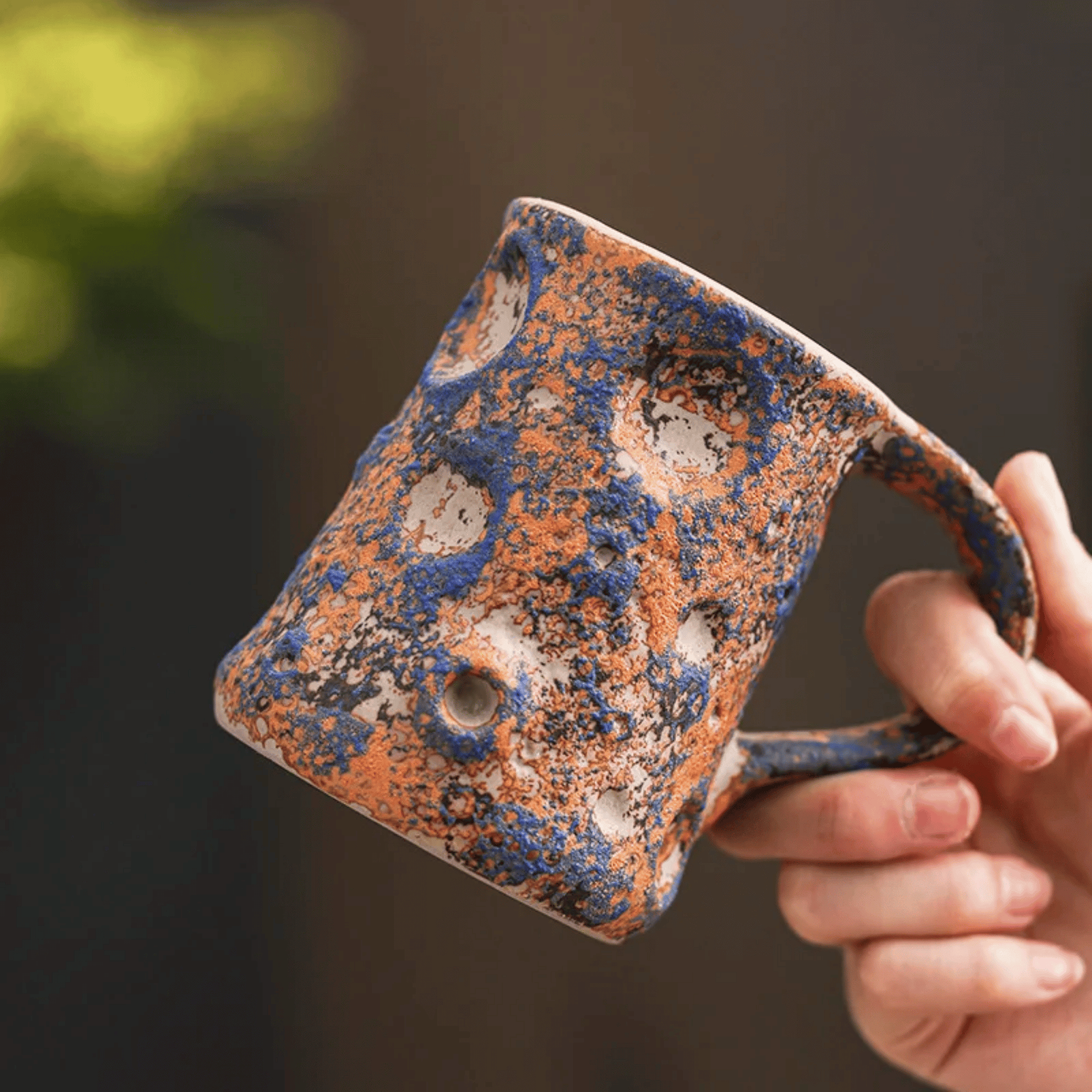 Hand holding a ceramic mug with blue and orange glaze against a blurred background