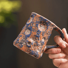Hand holding a ceramic mug with blue and orange glaze against a blurred background