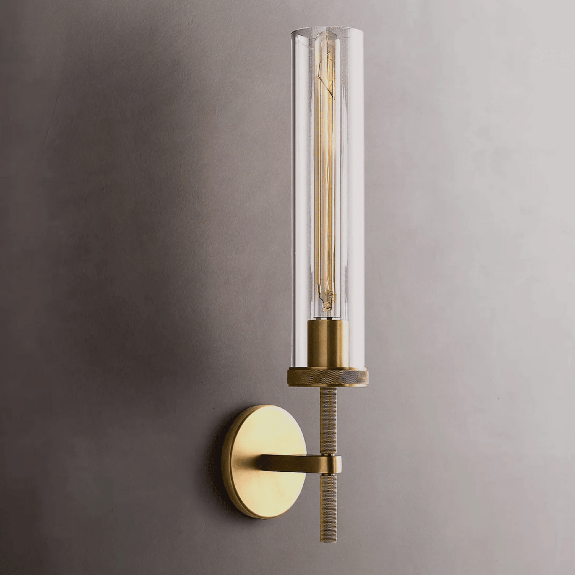 Wall-mounted light fixture with brass finish and clear glass on a gray wall.