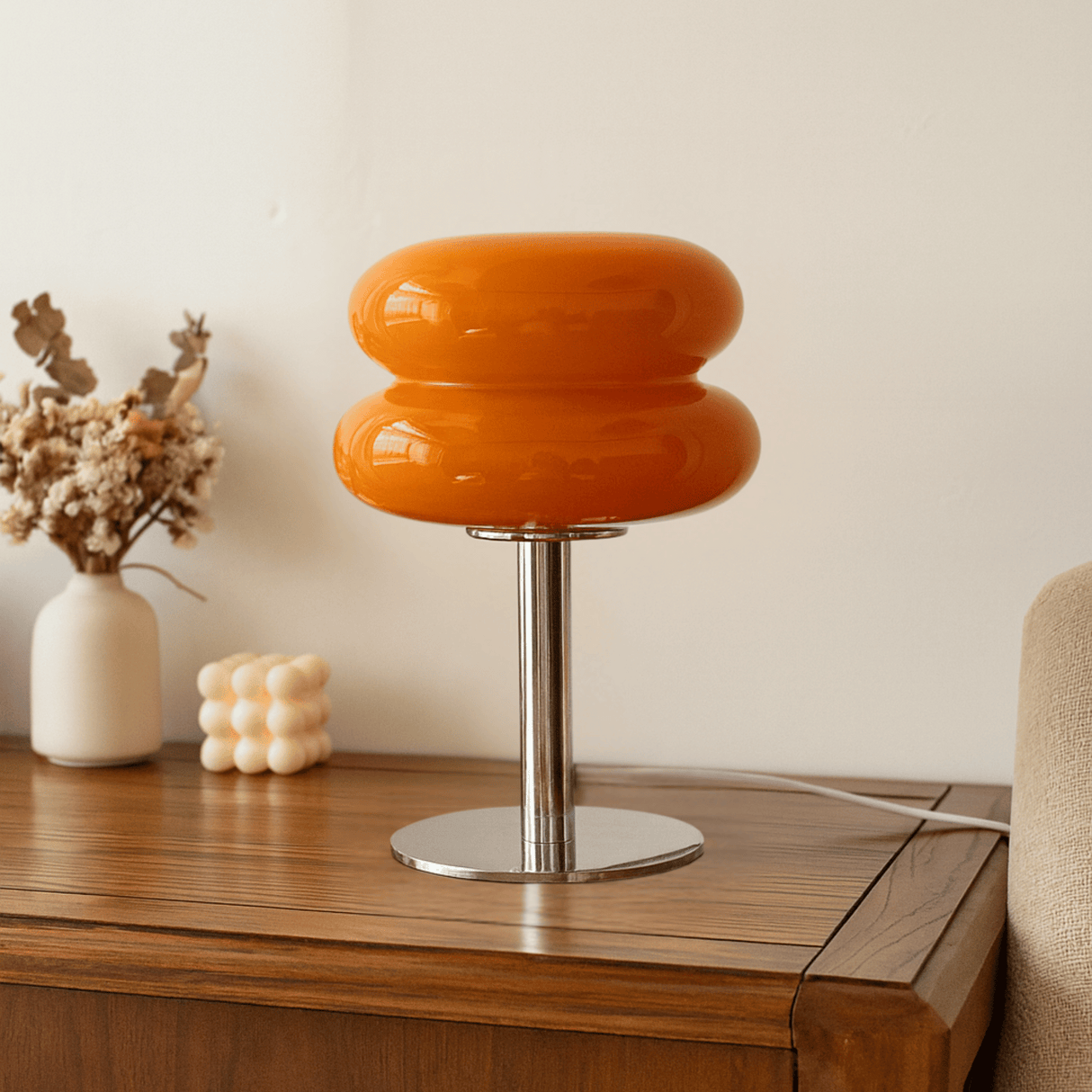 Orange table lamp with a metallic base on a wooden surface, with a vase and decorative items in the background.