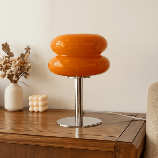 Orange table lamp with a metallic base on a wooden surface, with a vase and decorative items in the background.