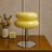 Yellow stool with a metallic base on a wooden surface, with decorative items in the background.