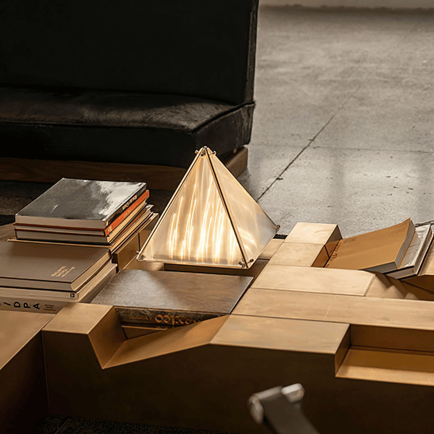 Modern wooden coffee table with geometric design, books, and a pyramid-shaped lamp in a dimly lit room.