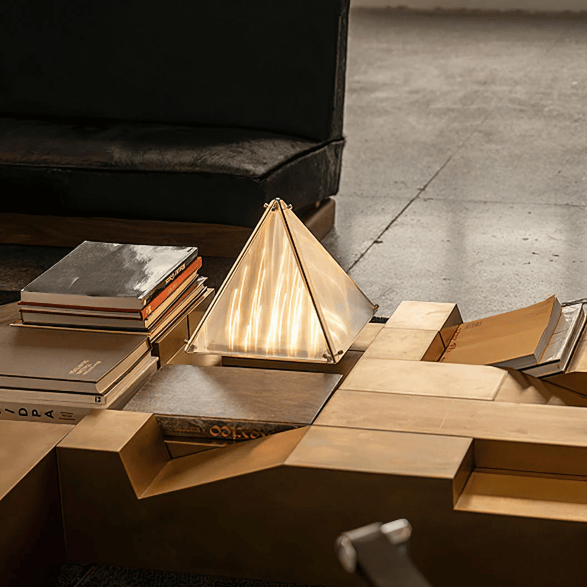 Modern wooden coffee table with geometric design, books, and a pyramid-shaped lamp in a dimly lit room.