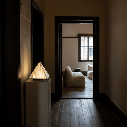 Dark hallway with a lamp on a pedestal and a room with a sofa and window in the background