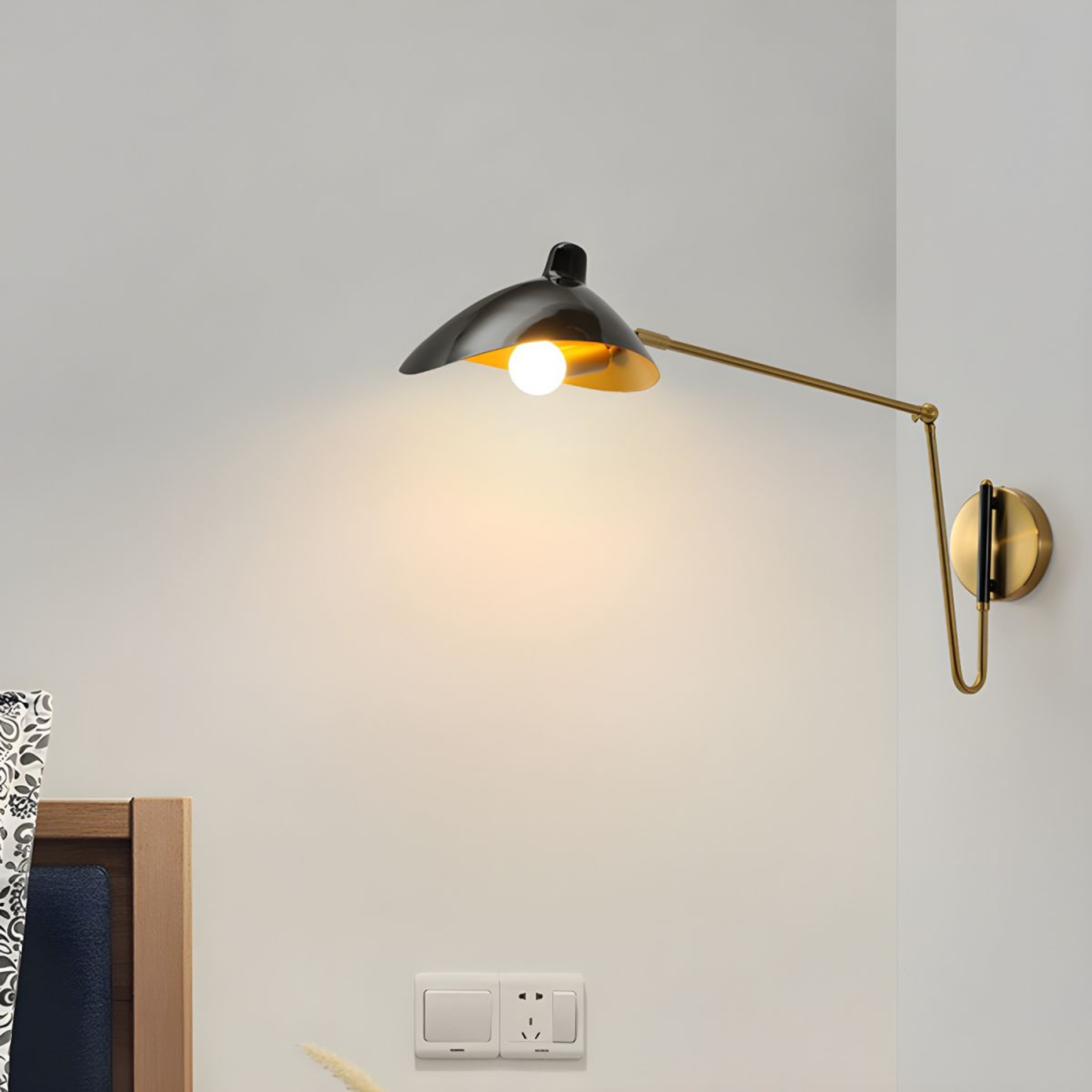 Modern black and gold adjustable wall lamp extending over a bedside area, casting warm light on a neutral wall.