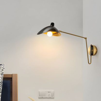 Modern black and gold adjustable wall lamp extending over a bedside area, casting warm light on a neutral wall.