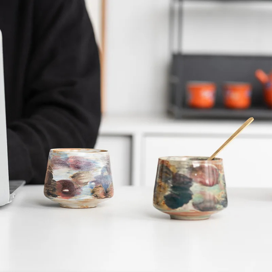 Close-up of two multicolor ceramic cups on a desk, one filled with coffee and being stirred with a small gold spoon.
