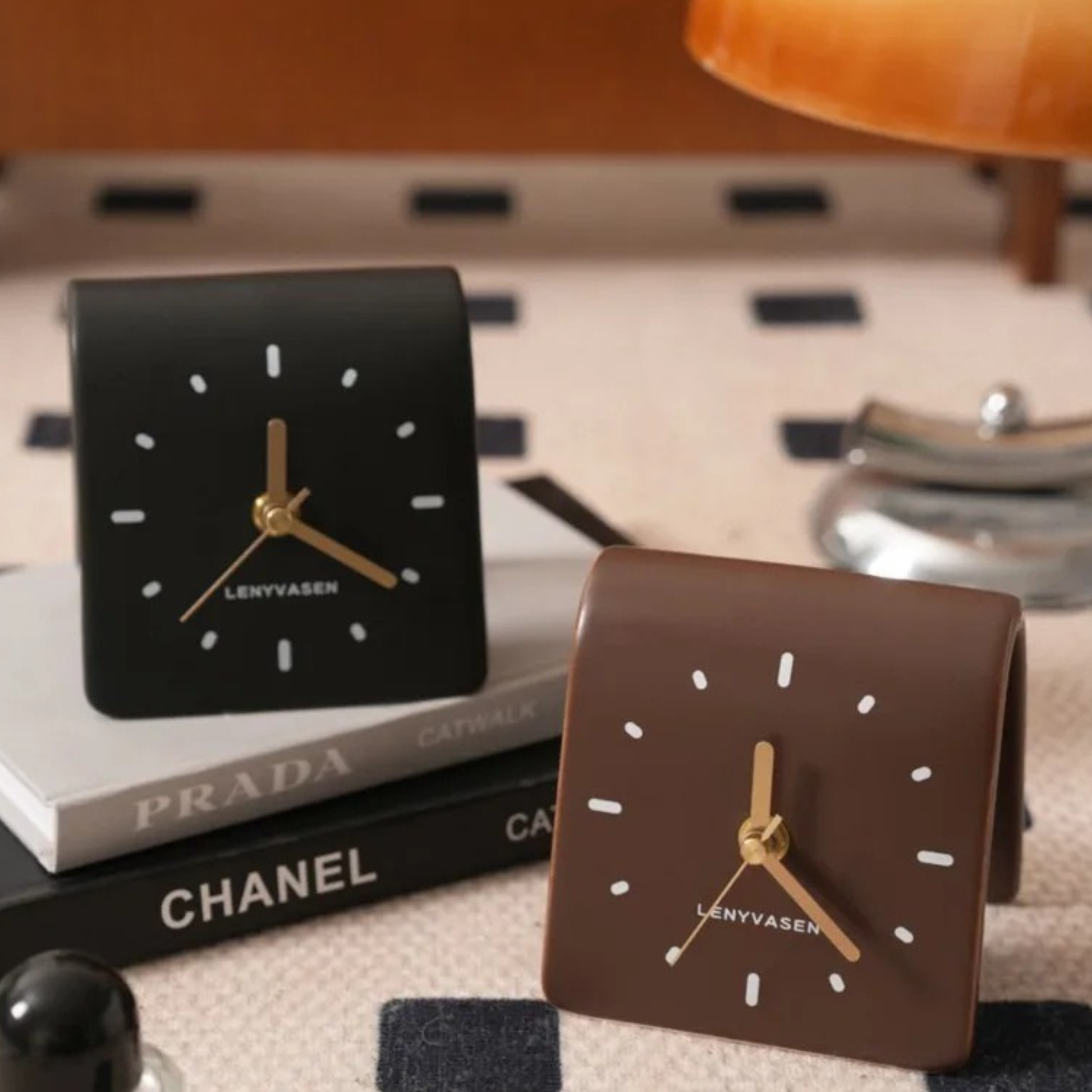 A brown leather desk clock with golden hands and square dial placed on a white desk beside books and a metallic decor piece.