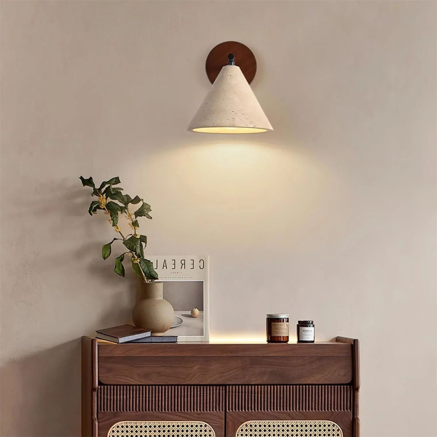 A wooden and white cone wall lamp mounted above a small wooden dresser with plants and decor, emitting soft warm light.