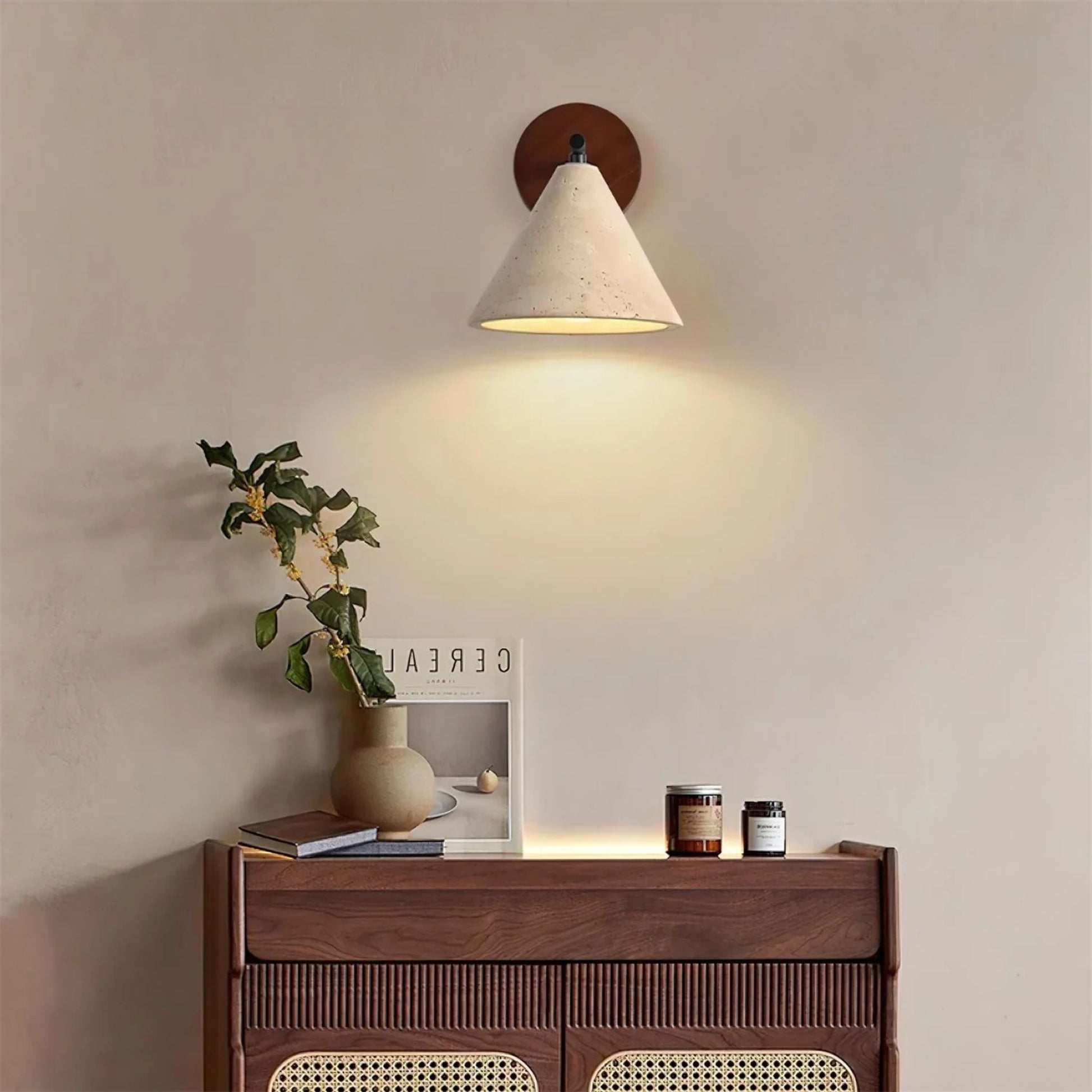 A wooden and white cone wall lamp mounted above a small wooden dresser with plants and decor, emitting soft warm light.