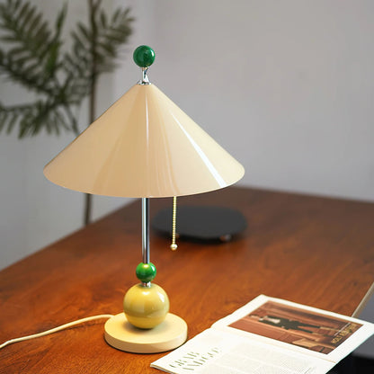 A unique table lamp with a beige cone-shaped shade and green accent balls, placed on a wooden table with a few books.