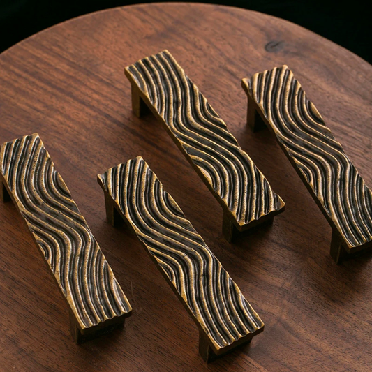 Four rectangular brass drawer handles with wavy engraved patterns placed on a wooden surface.