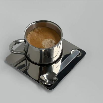 Stainless steel coffee cup on a reflective square saucer with spoon, set on a wooden table beside an open book and candle.