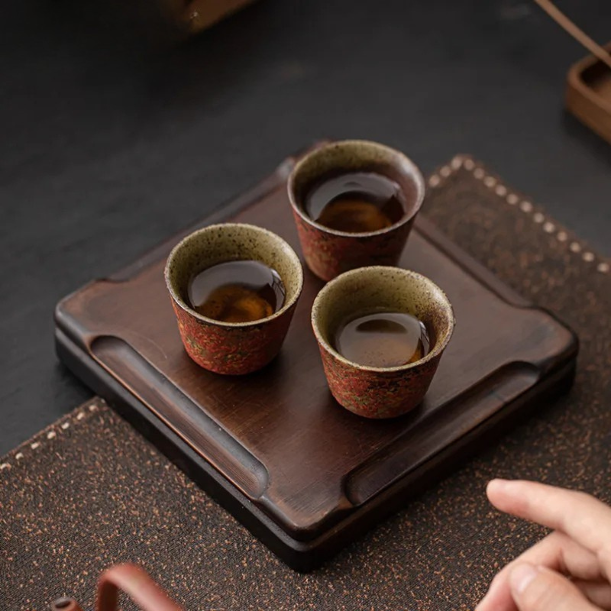 Three small red ceramic tea cups filled with tea, placed on a wooden tray on a tea table.