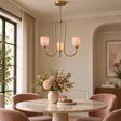 A modern three-light brass chandelier with frosted pink glass shades hangs over a round marble dining table in a softly styled dining room with blush pink chairs and floral centerpieces.