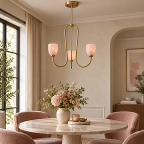 A modern three-light brass chandelier with frosted pink glass shades hangs over a round marble dining table in a softly styled dining room with blush pink chairs and floral centerpieces.