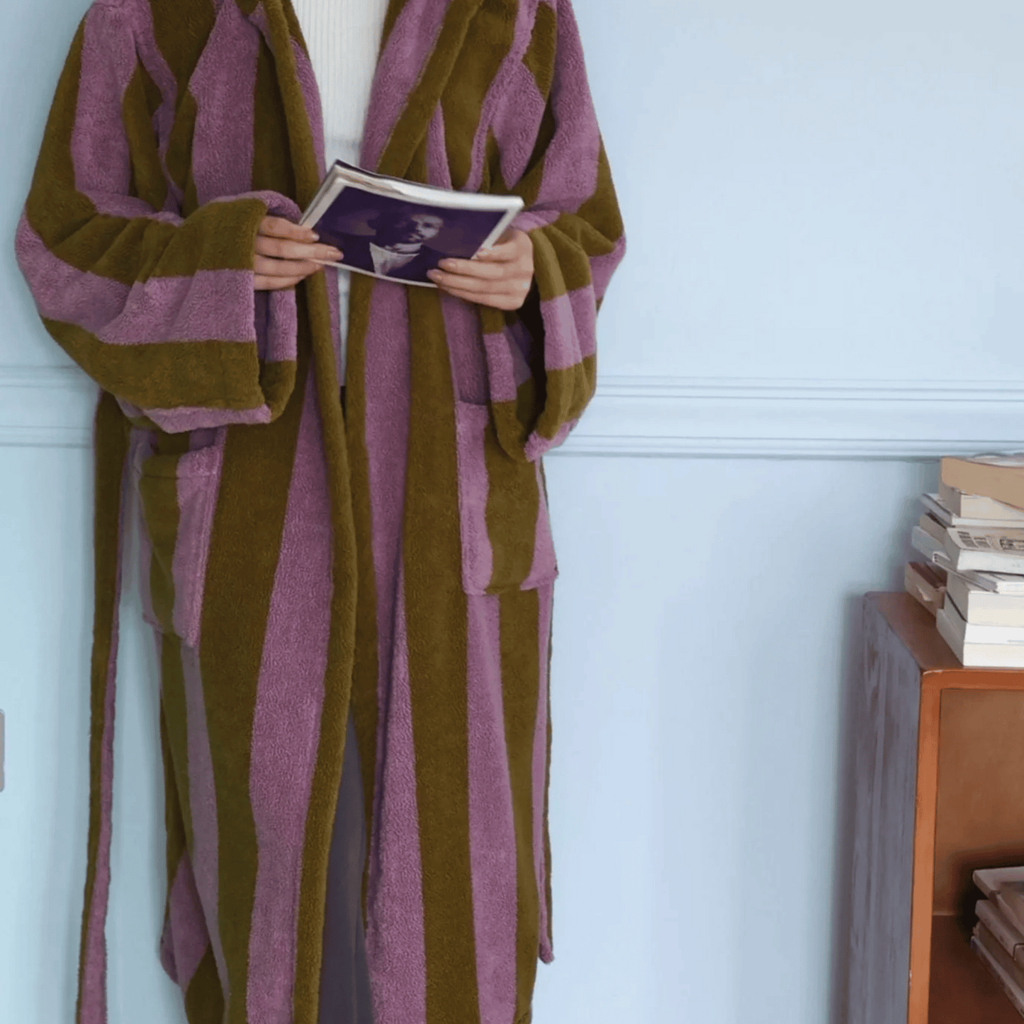 Person wearing a brown and purple striped robe holding a book against a light blue wall.