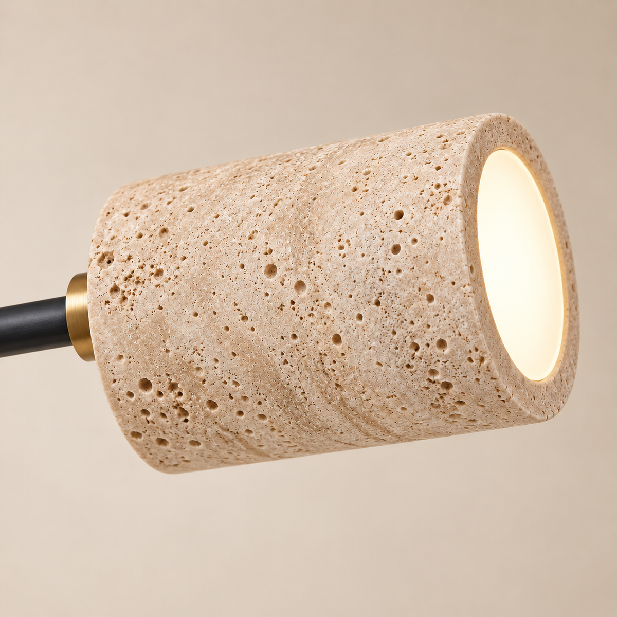 A modern cylindrical pendant light with a textured beige travertine stone shade, a black metal stem, and a gold accent collar, shown illuminated against a warm beige background.