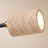 A modern cylindrical pendant light with a textured beige travertine stone shade, a black metal stem, and a gold accent collar, shown illuminated against a warm beige background.