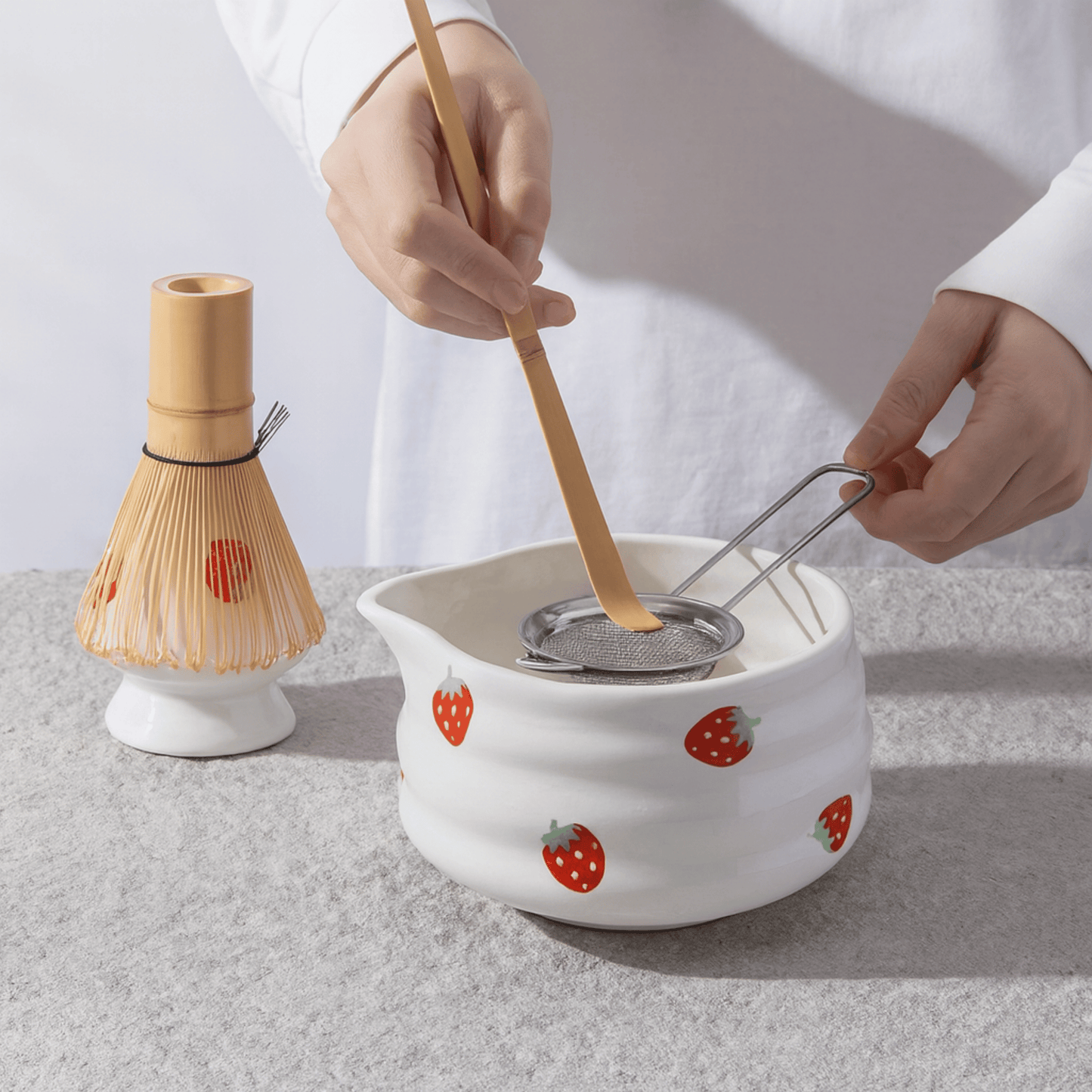 Person using a wooden whisk and strainer over a white ceramic pot with strawberry design on a light gray background.