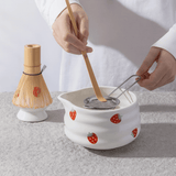 Person using a wooden whisk and strainer over a white ceramic pot with strawberry design on a light gray background.