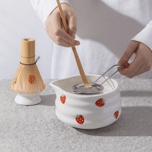 Person using a wooden whisk and strainer over a white ceramic pot with strawberry design on a light gray background.