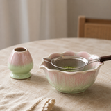 A ceramic matcha whisk bowl with a scalloped rim, shown alongside a small matching pink and green ceramic scoop on a beige tablecloth. A metal sieve rests inside the bowl.