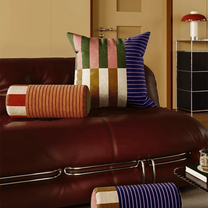 Decorative pillows on a brown leather sofa with a lamp and cabinet in the background.