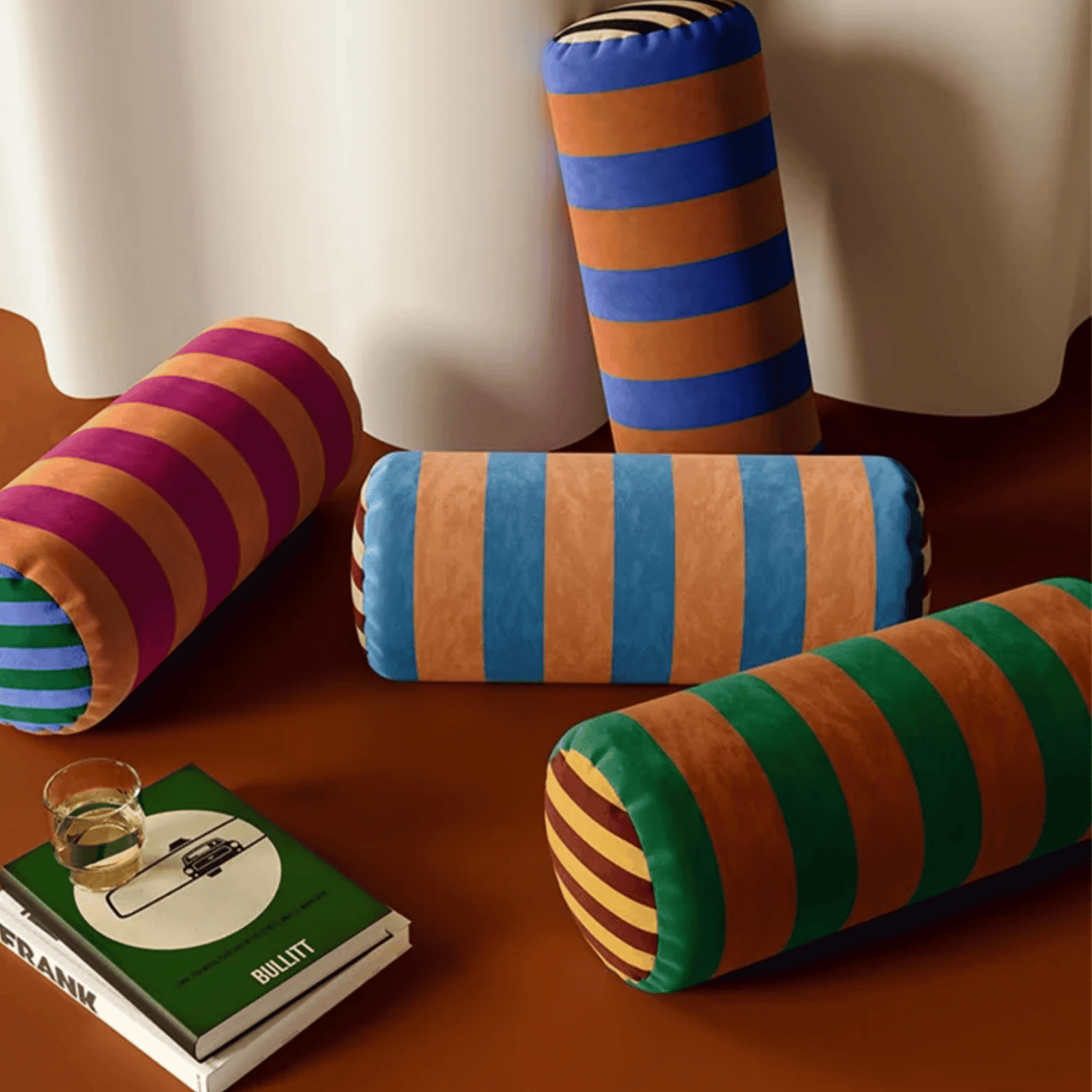 Colorful striped cylindrical pillows on a wooden surface with a book and glass.