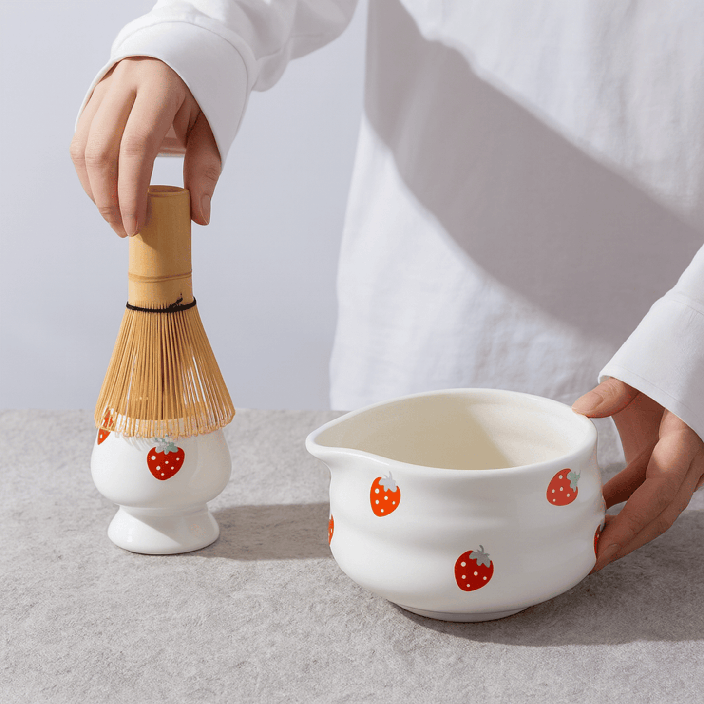Person holding a white ceramic bowl with strawberry pattern and a bamboo whisk on a light gray background