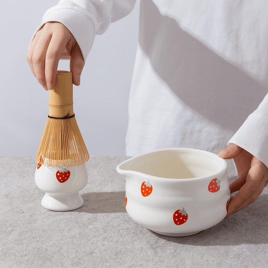 Person holding a white ceramic bowl with strawberry pattern and a bamboo whisk on a light gray background