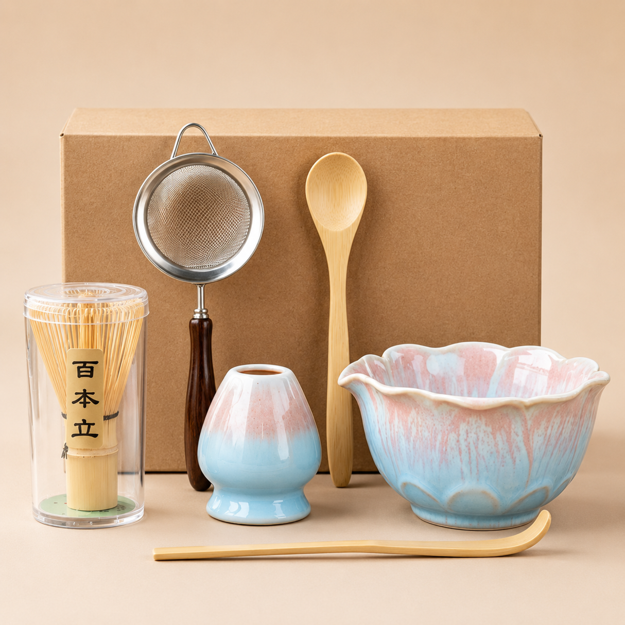 A Japanese matcha tea set displayed in front of a brown kraft box, including a pastel blue and pink ceramic bowl, a small matching cup, a wooden spoon, a metal strainer, a bamboo whisk in a clear container, and a wooden scoop.