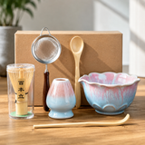 Ceramic tea set on a wooden table with a brown gift box behind it, including a pink-and-blue glazed bowl, small matching cup, bamboo whisk in a clear container, metal strainer, wooden spoon, and chopsticks.