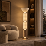 A modern multi-tiered floor lamp with a white bowl-shaped top, three glowing cylindrical shades, and a light wood base, standing in a warmly lit living room next to a curved beige sofa.
