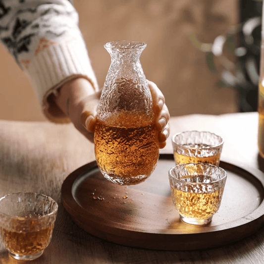 Person pouring liquid from a glass carafe into small glasses on a wooden tray.