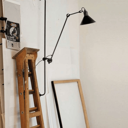 Black wall lamp with long arm on a white wall next to a wooden ladder and framed picture.