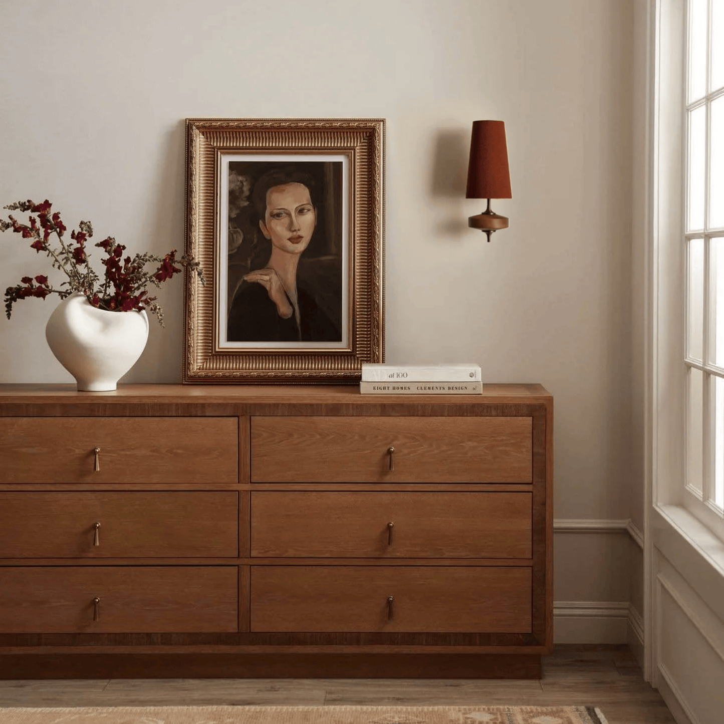 Wooden dresser with a framed portrait, vase, and books in a room with a lamp and window.