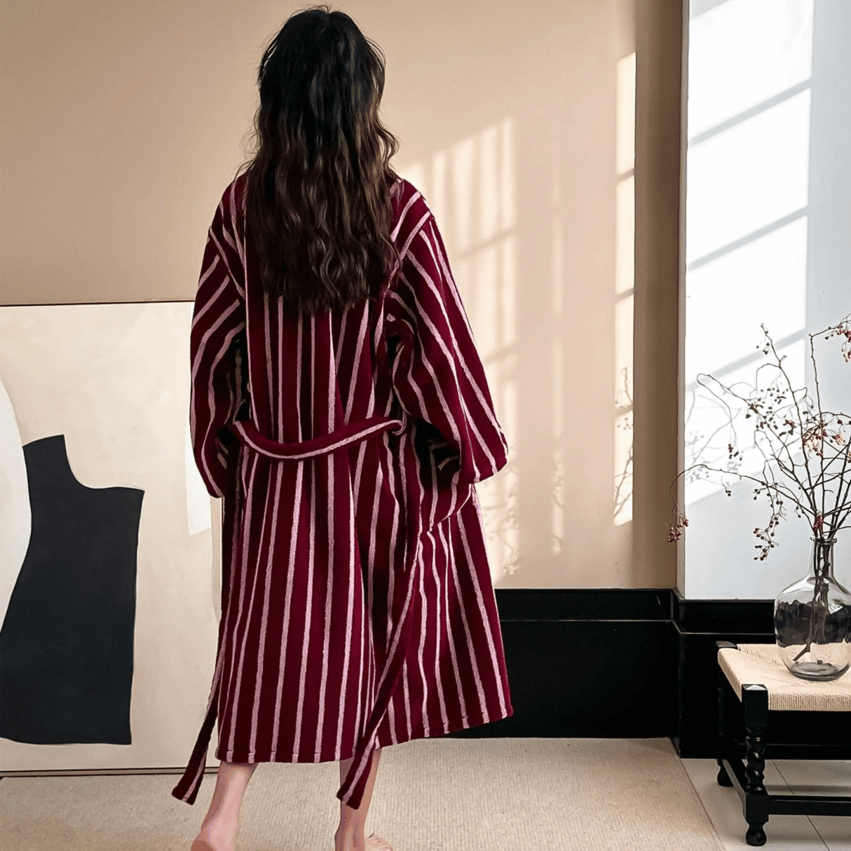 Woman wearing a burgundy and white striped robe in a room with a window and black coat on a stand.