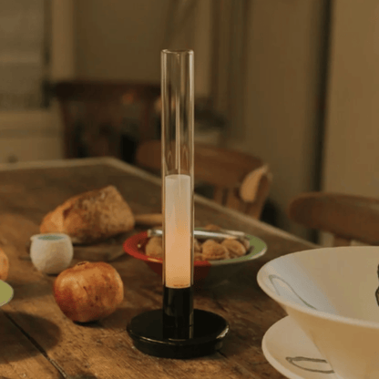 Candle in a cylindrical holder on a wooden table with food items in the background