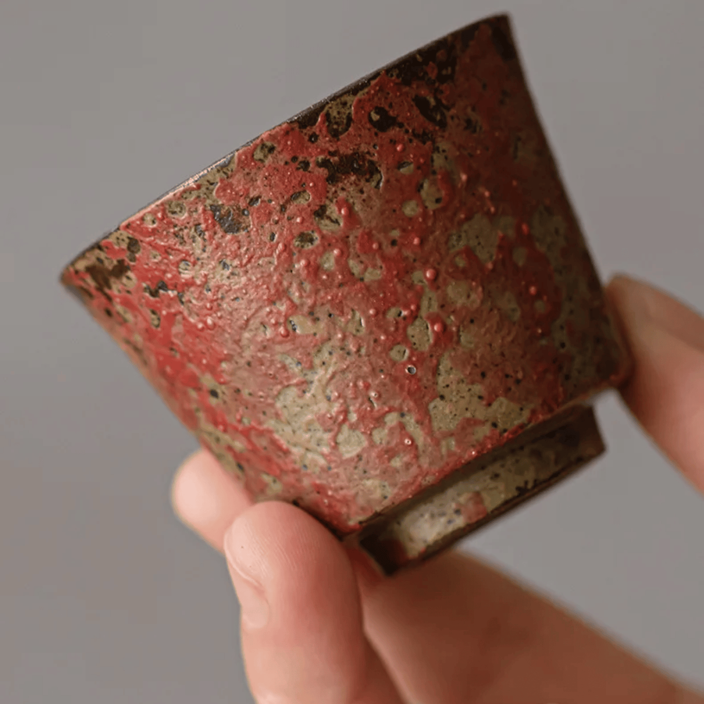 Hand holding a square ceramic cup with red and brown glaze against a neutral background