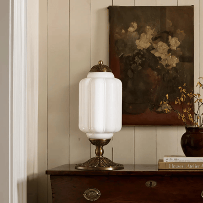 Decorative table lamp with white glass shade and gold base on a wooden surface, with a painting of flowers in the background.