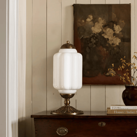 Decorative table lamp with white glass shade and gold base on a wooden surface, with a painting of flowers in the background.