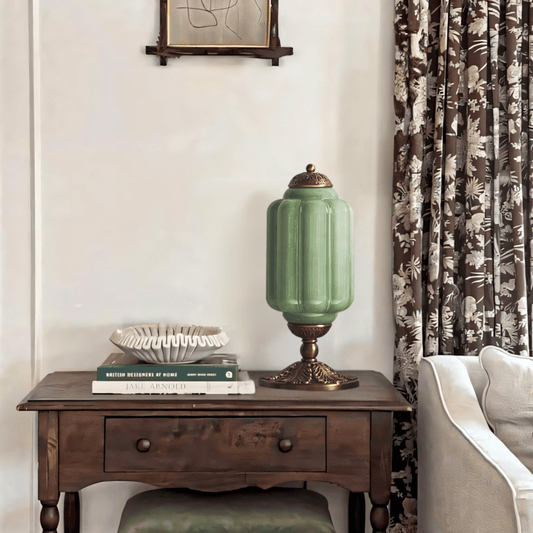 Green lamp on a wooden side table with books and a decorative bowl, next to a floral curtain.