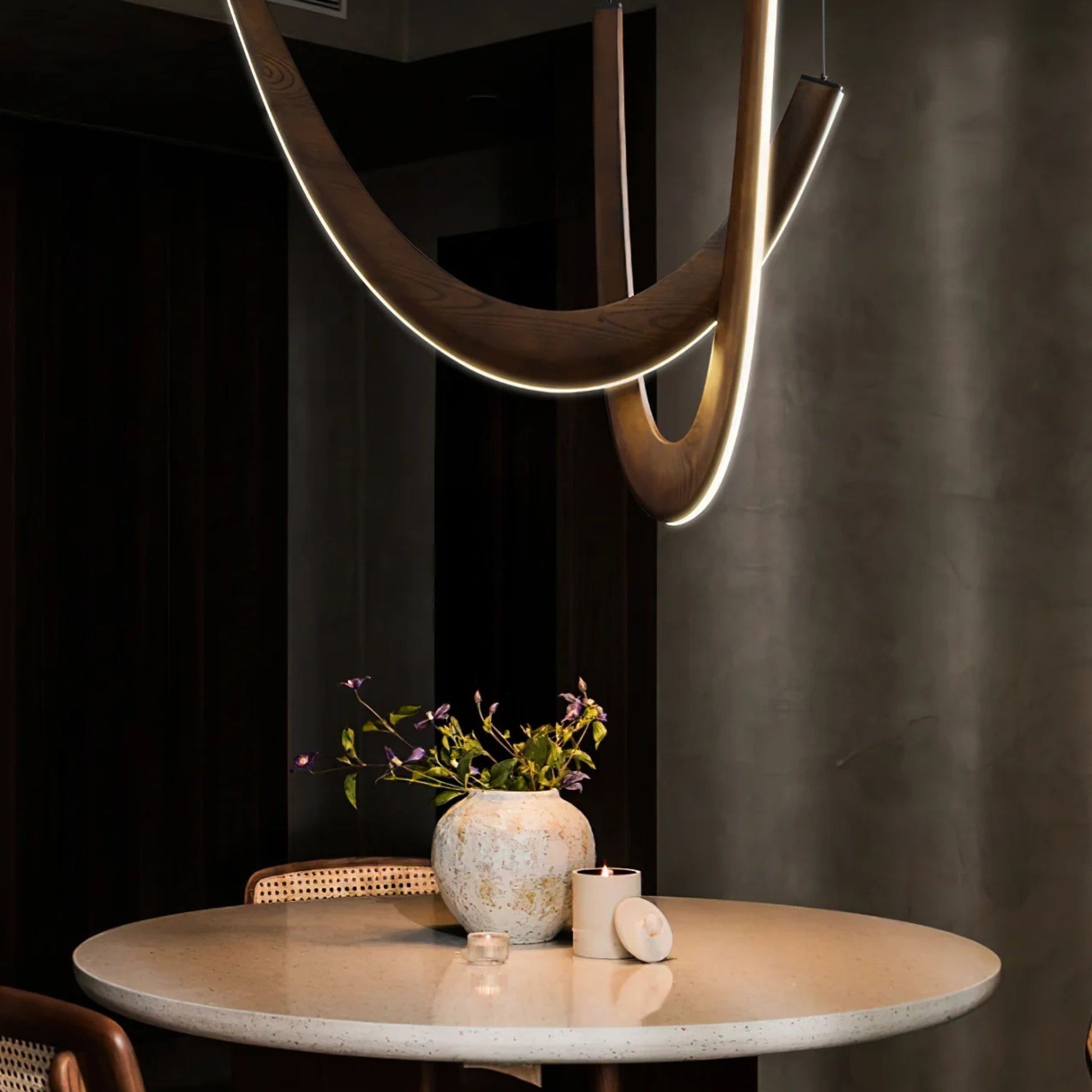A dimly lit dining room with a contemporary pendant light featuring curved wooden arms, hanging above a round table.