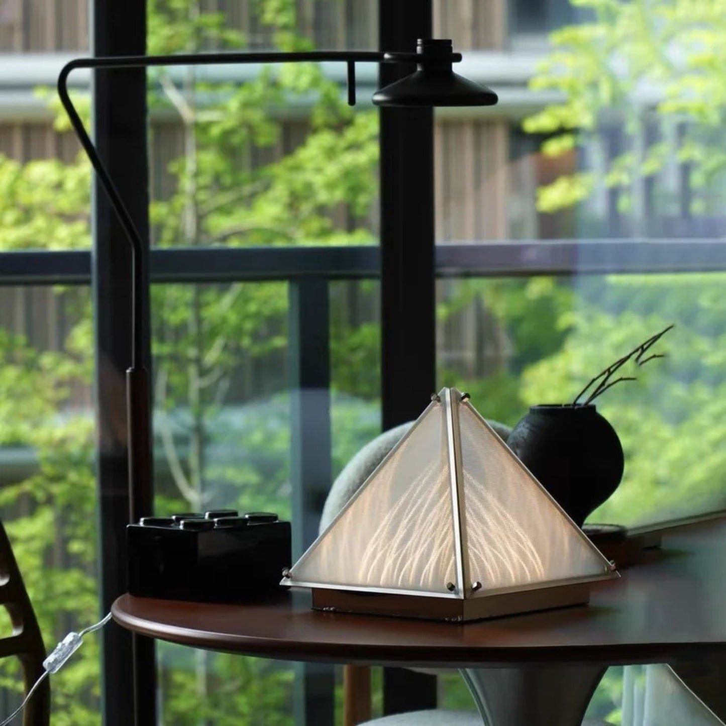 A glowing cone-shaped lamp on a round wooden table in front of large windows overlooking greenery.