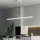 A minimalist contemporary kitchen featuring white cabinetry, a wooden open shelf, a compact white microwave, and two rattan chairs set around a small dining table illuminated by a stylish black pendant light.