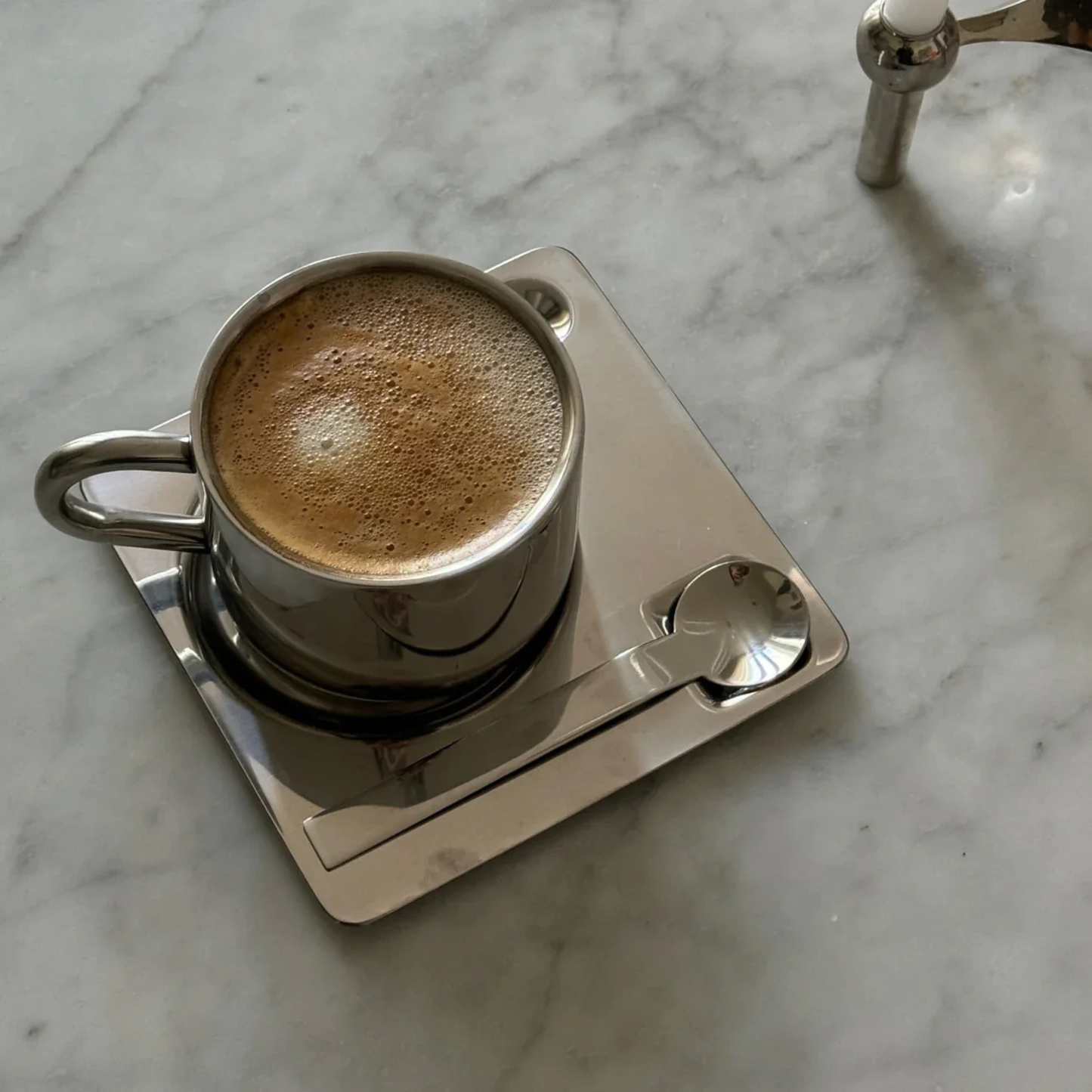 Top view of a stainless steel coffee cup filled with foam coffee on a reflective square saucer with a metal spoon.